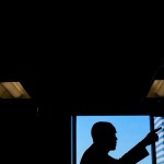 A man in silhouette points into an unseen crowd, backlit against a window showing blue sky.