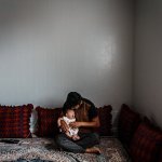 A father holds his newborn baby in his lap while seated on the floor.