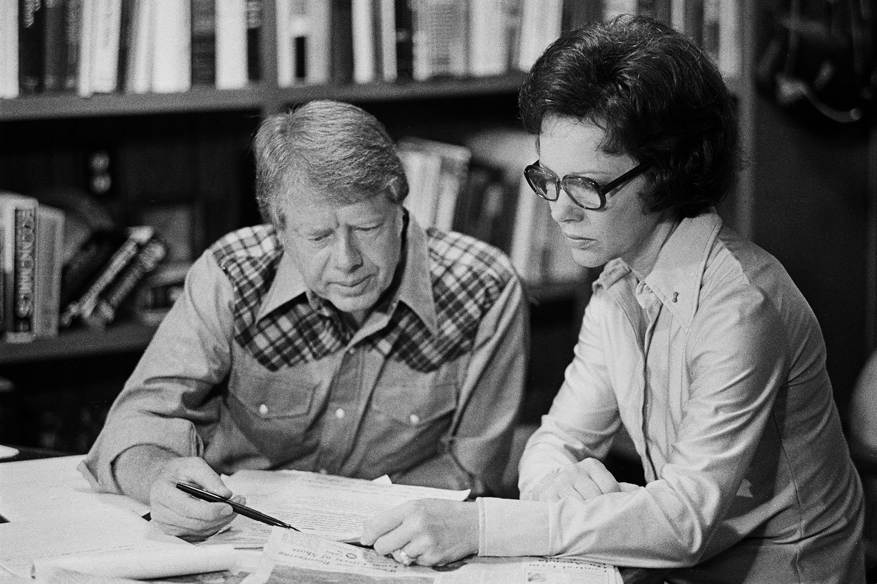 Jimmy Carter, wearing a shirt with plaid patches on the shoulders, and his wife, Rosalynn Carter, reading a document on the table before them.