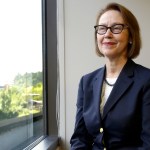 A woman in a blue suit smiles as she looks out a window with an American flag in the foreground.