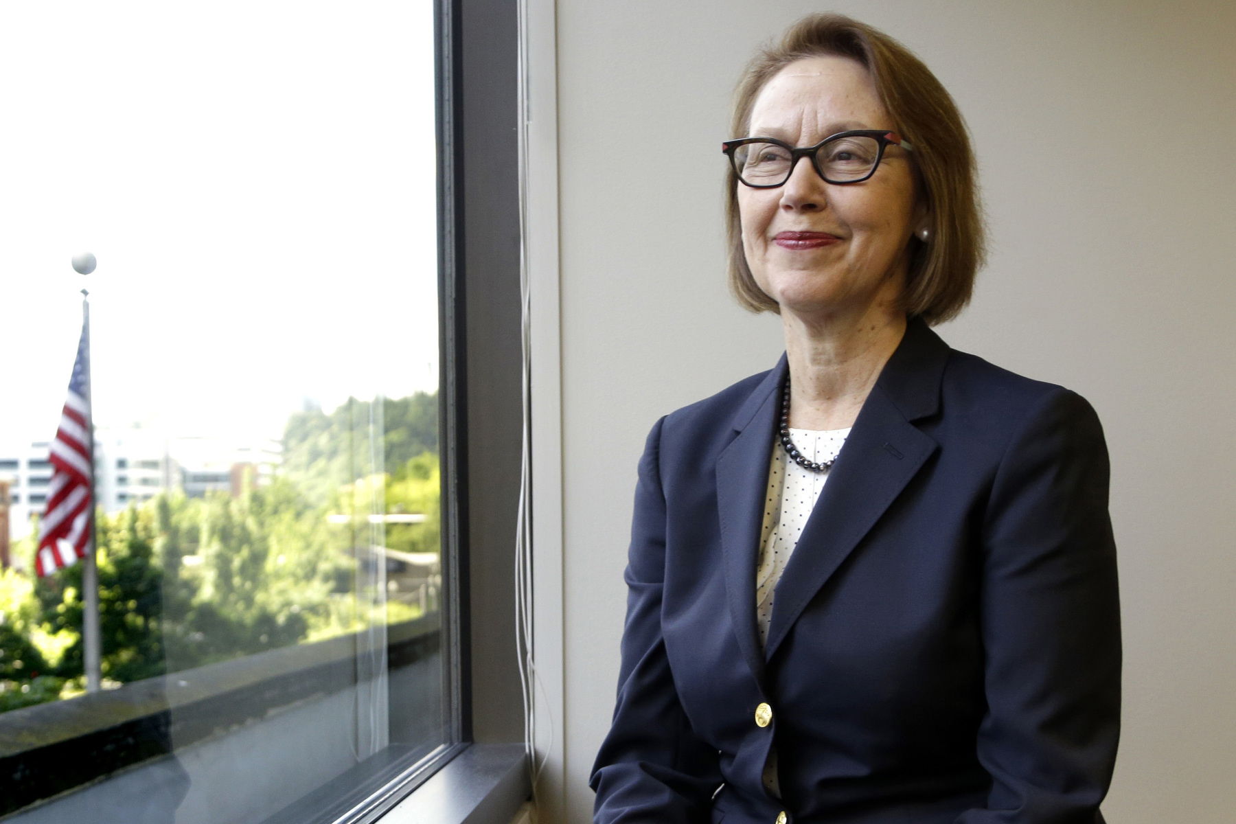 A woman in a blue suit smiles as she looks out a window with an American flag in the foreground.