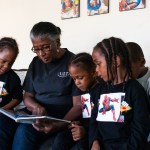 Four children sit on a sofa listening to a woman reading them a book.