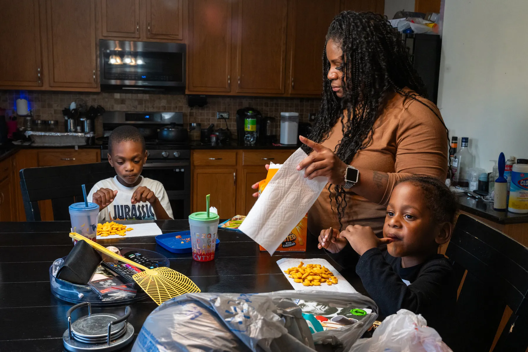 Two of the triplets sit at a dining table while their mother stands beside them, making them a snack.