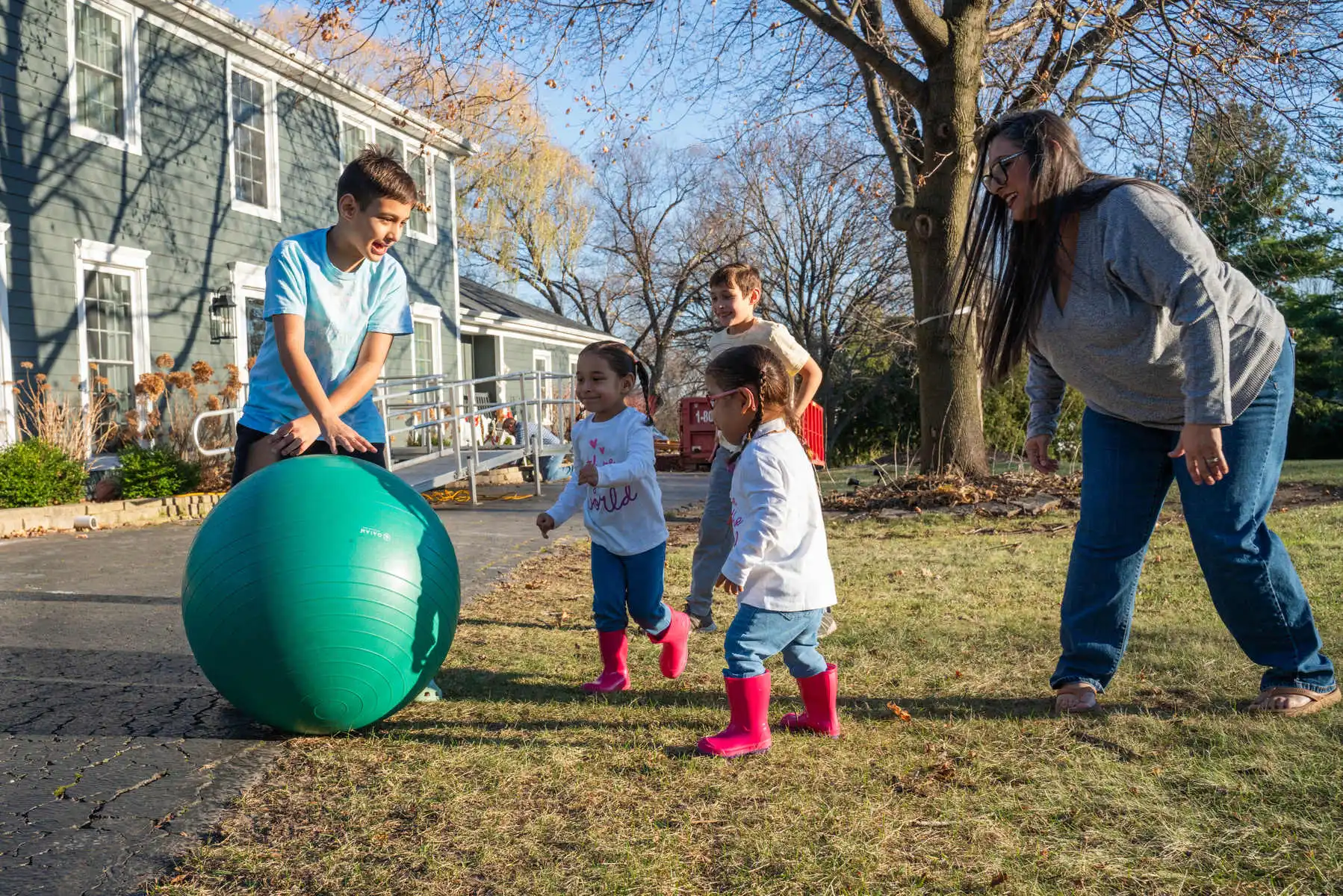 Four children run after a big ball outside as their mother plays with them.