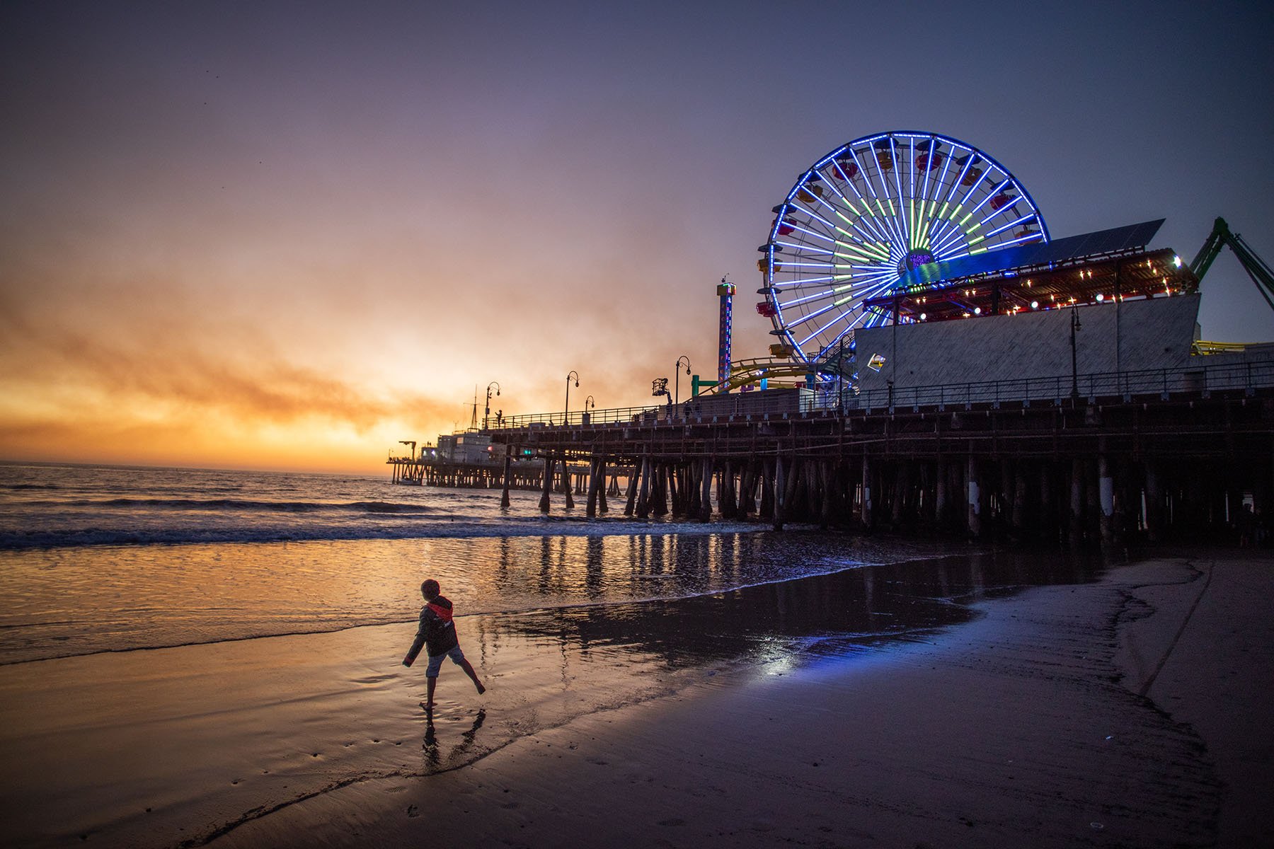 A boy plays on the beach with the Santa Monica pier on the background with smoke from the Palisades Fire seen in the sky.