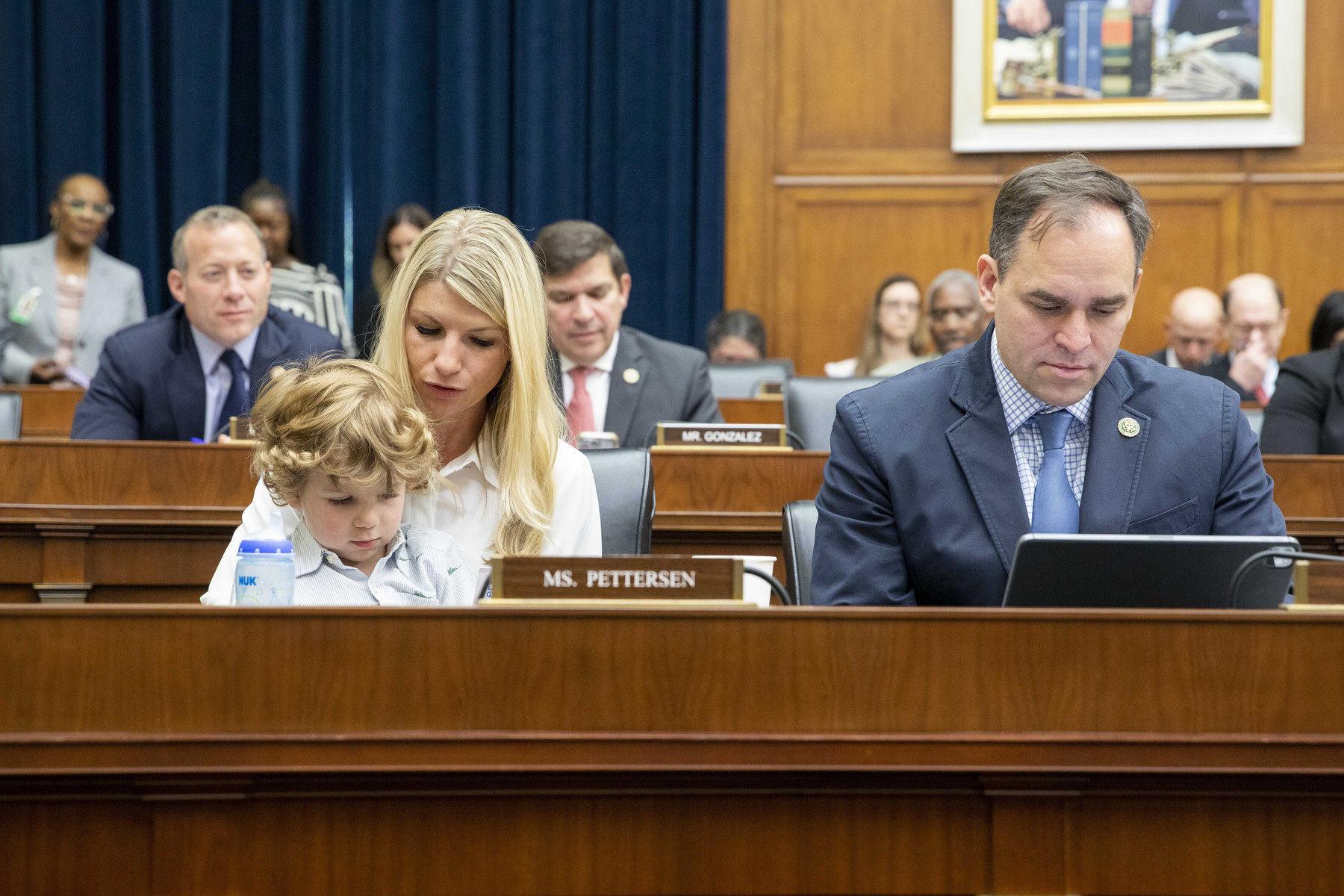 A woman sits with a toddler in her lap beside a man in a room of politicians.