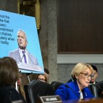 A woman speaks from behind a raised desk with a poster behind her of RFK Jr. and his previous statements on women's bodily autonomy.