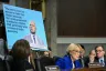 A woman speaks from behind a raised desk with a poster behind her of RFK Jr. and his previous statements on women's bodily autonomy.