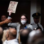 A protestor holding a NO RFK Jr sign is grabbed by a security guard in a room full of people.