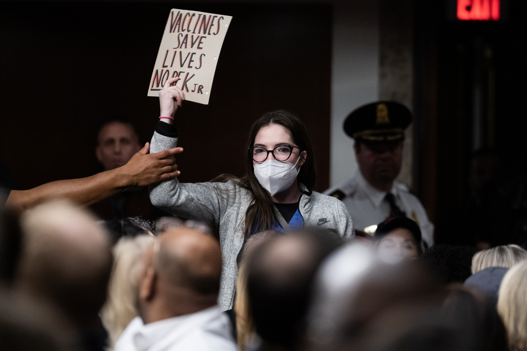 A protestor holding a NO RFK Jr sign is grabbed by a security guard in a room full of people.