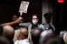 A protestor holding a NO RFK Jr sign is grabbed by a security guard in a room full of people.