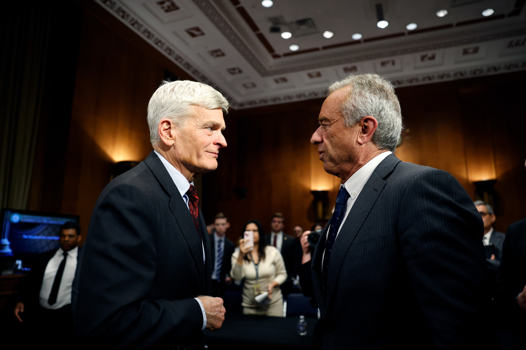 Two men speak to each other standing in a room full of people.