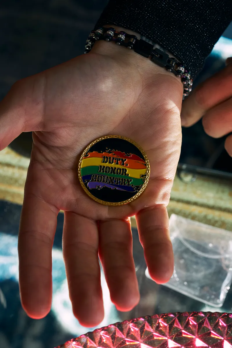 A hand holds a medallion that says duty, honor, country over a rainbow motif.