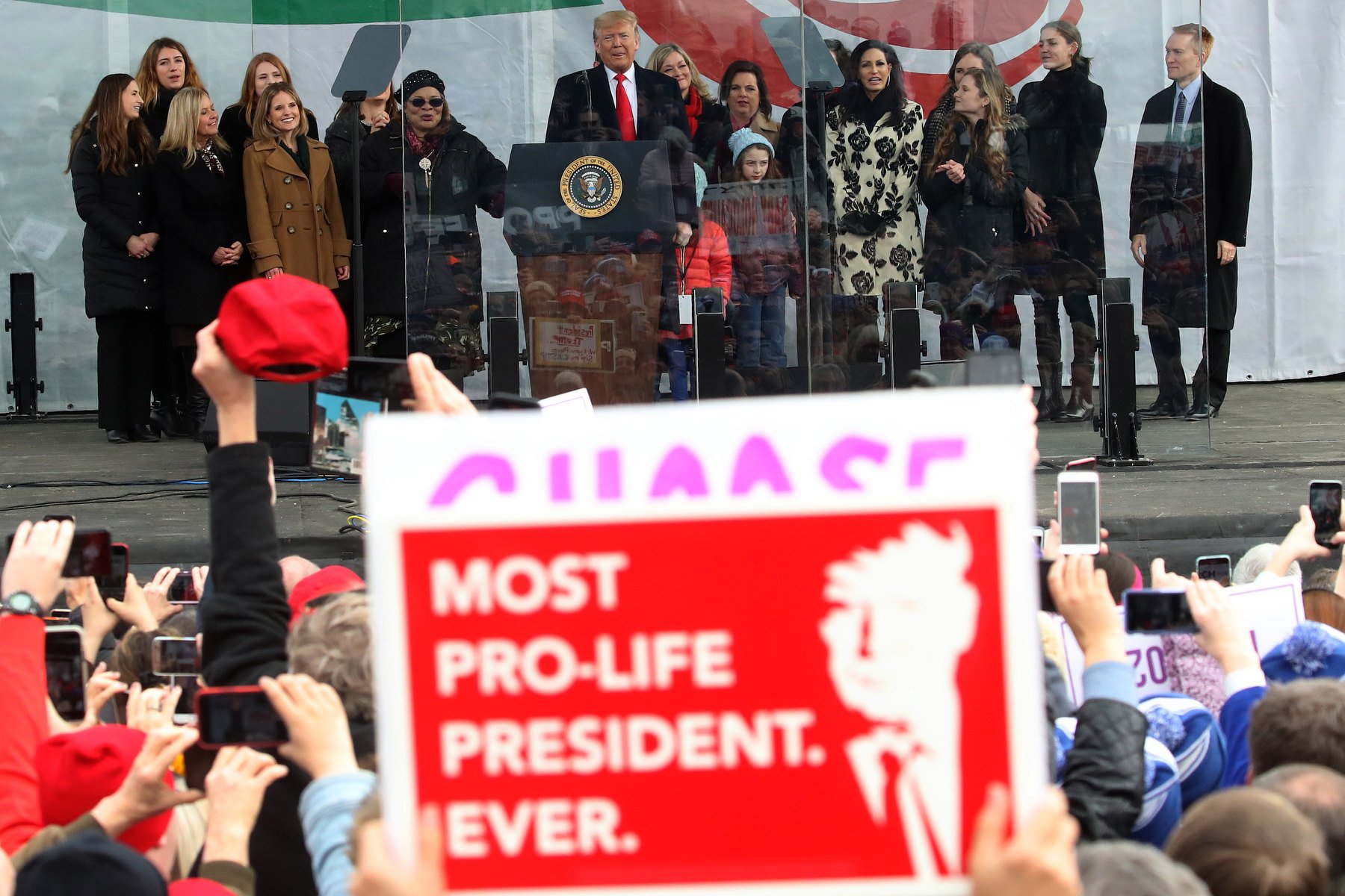 A sign saying "most pro-life president ever" is held up in a crowd watching Trump speak at a podium.