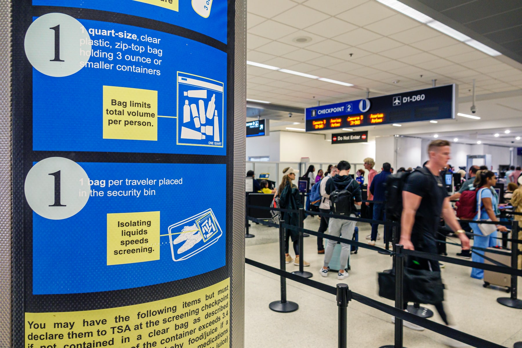 A TSA sign with limits on liquids is seen with a passenger security line in the background