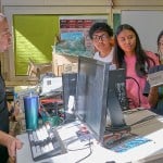 A teacher at Farrington High School instructs three students, one of whom is holding a laptop up to him for review.