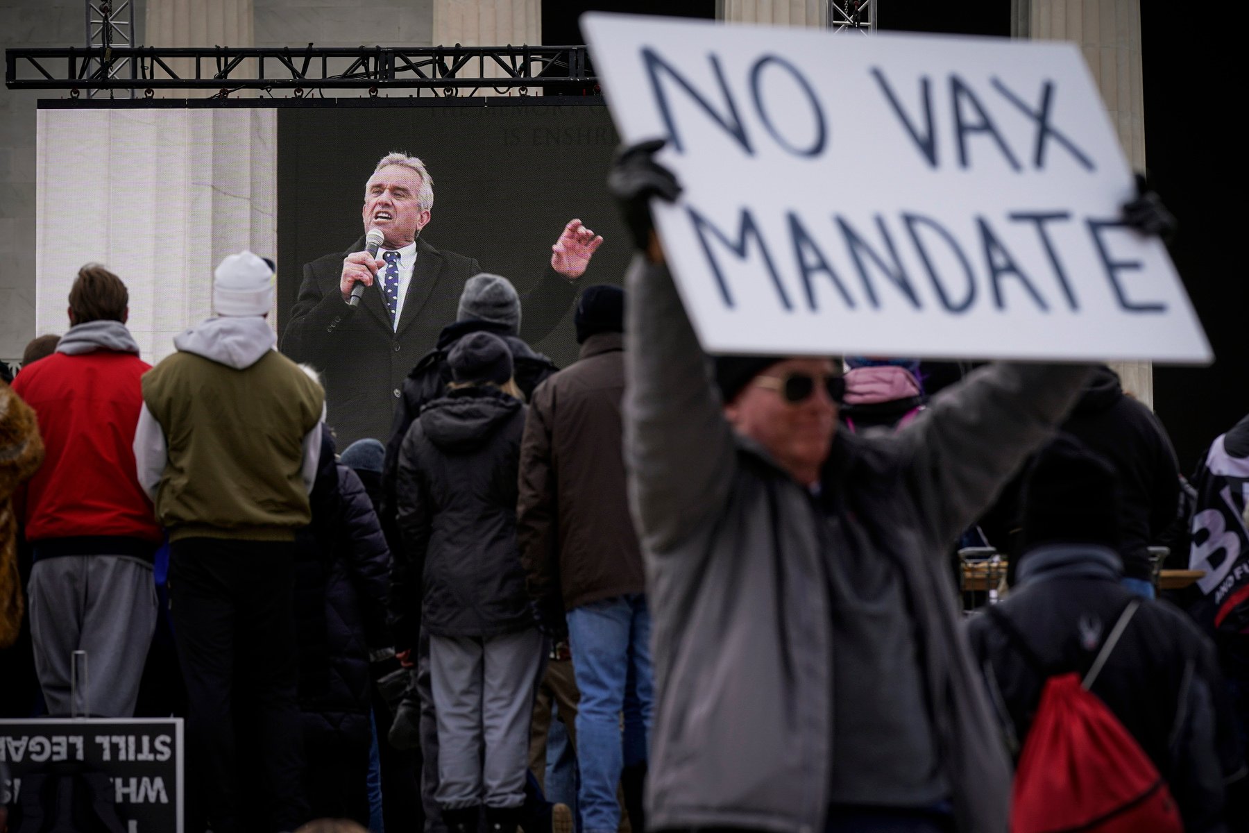 A man holds a sign that says "no vax mandate" while Kennedy speaks onstage in the background.