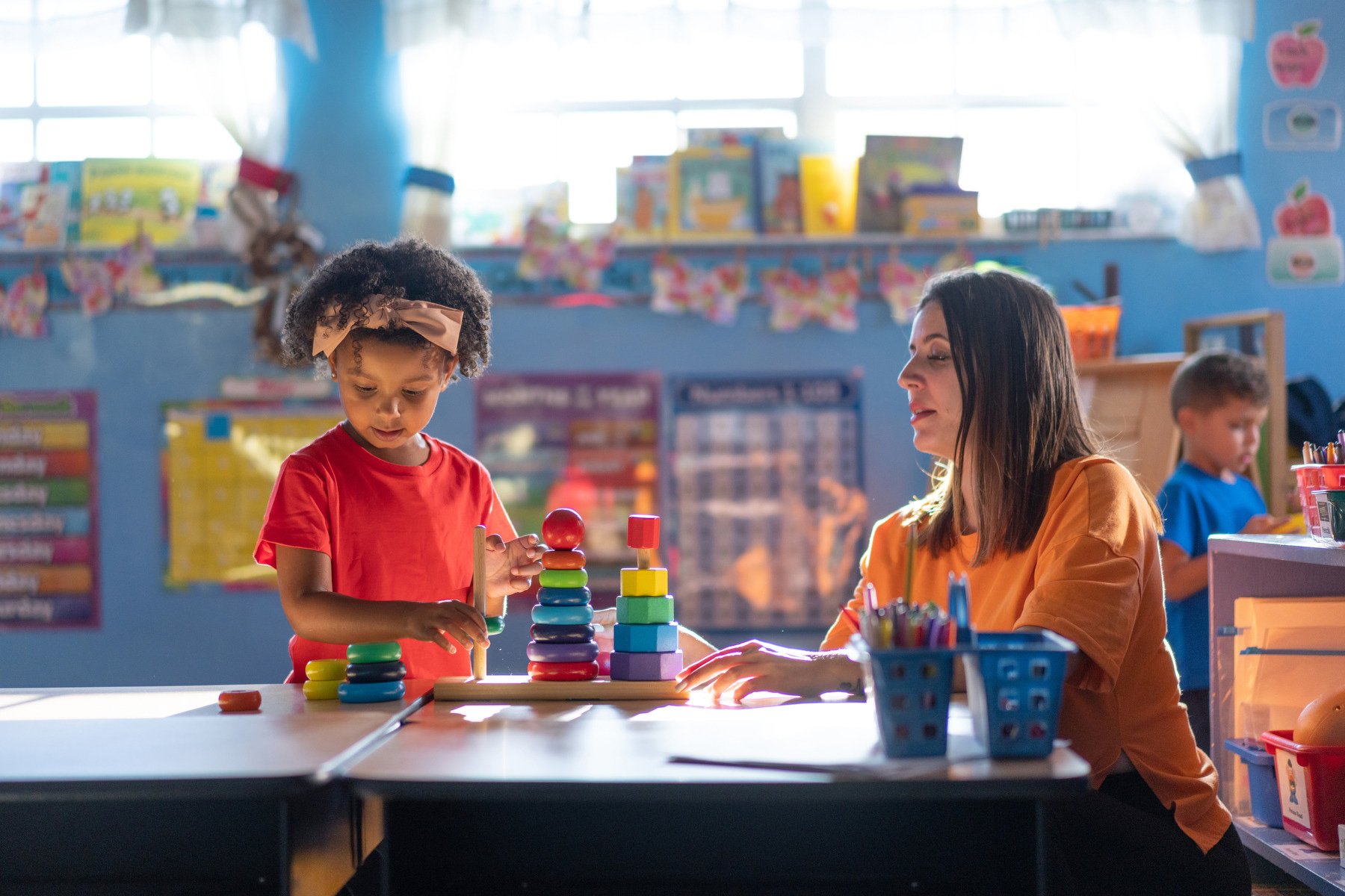 A preschooler plays with toys beside her teacher.