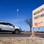 A white car drives past a sign pointing to a measles testing site.