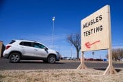 A white car drives past a sign pointing to a measles testing site.