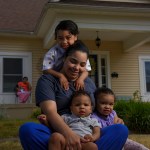 A mother holds her three children as she smiles in front of a home.