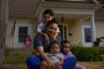 A mother holds her three children as she smiles in front of a home.