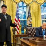 Elon Musk, wearing a black Make America Great Again cap, stands in the Oval Office next to President Donald Trump, who is seated at the Resolute Desk