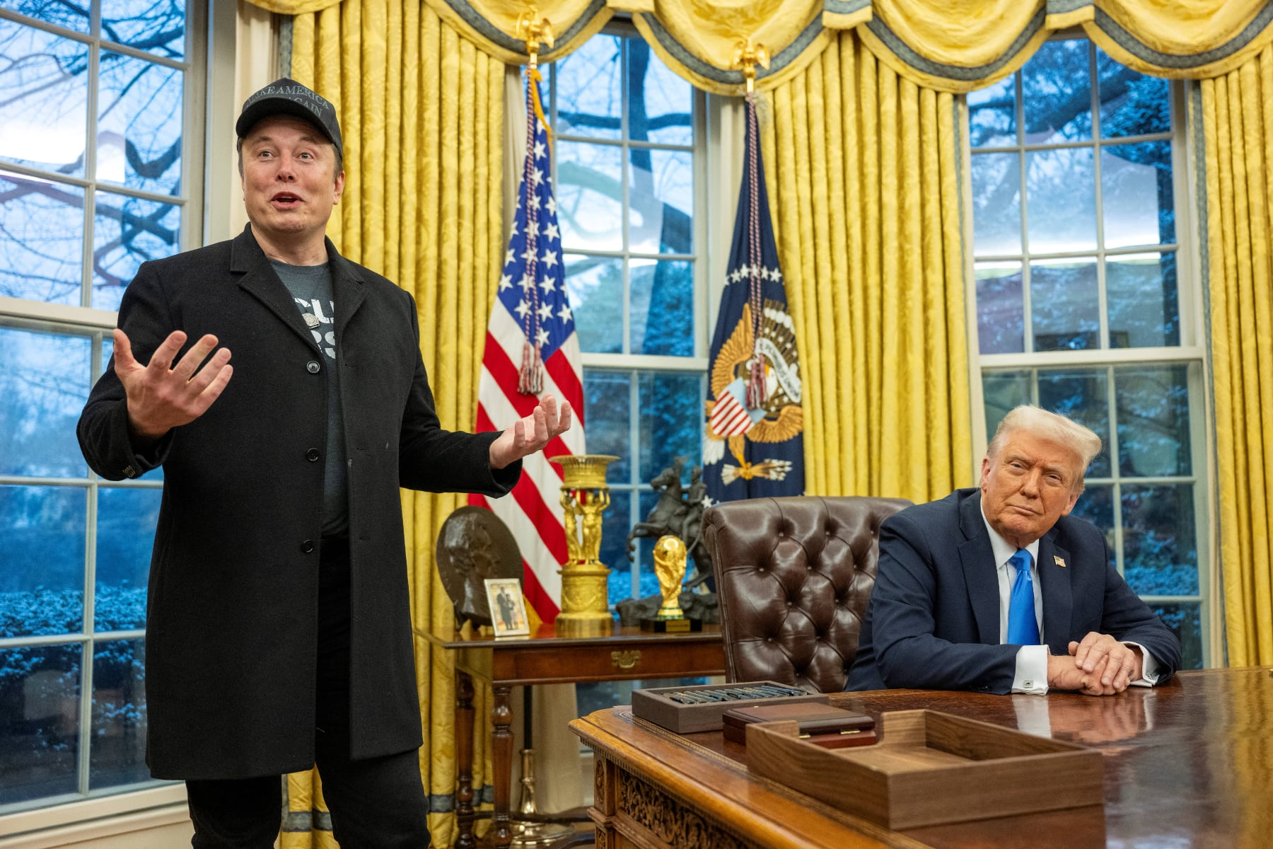 Elon Musk, wearing a black Make America Great Again cap, stands in the Oval Office next to President Donald Trump, who is seated at the Resolute Desk