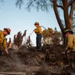 Firefighters spray water and use hoes to contain a barely visible fire on a property where a house formerly stood.