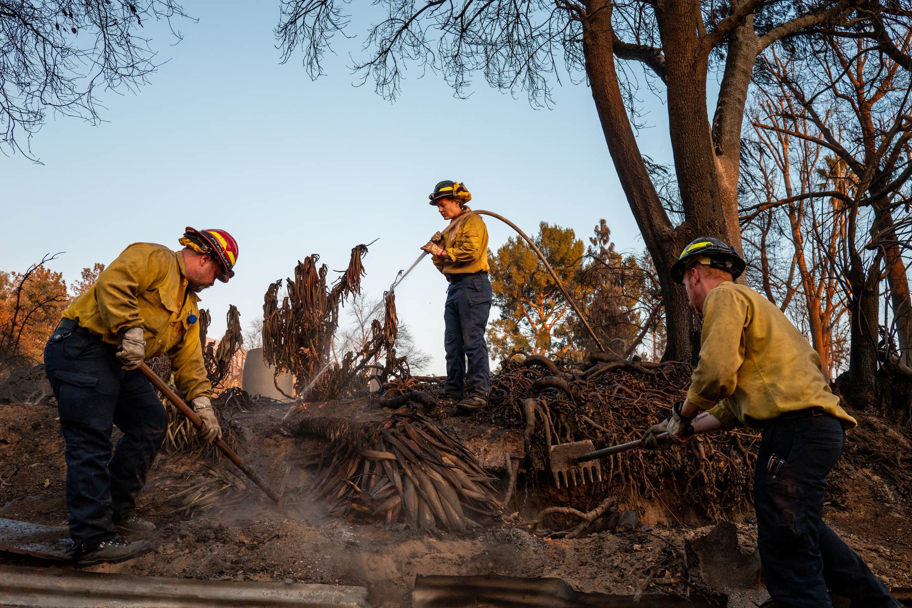 Firefighters spray water and use hoes to contain a barely visible fire on a property where a house formerly stood.