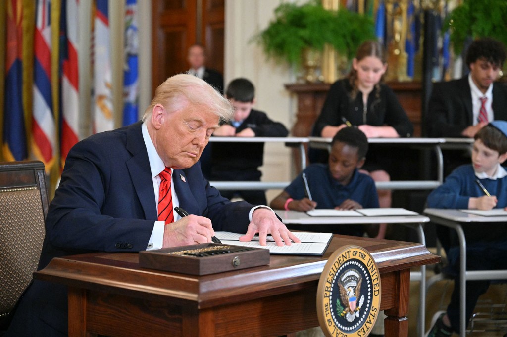 President Donald Trump signs an executive order surrounded by children at school desks also signing fake executive orders in the East Room of the White house.