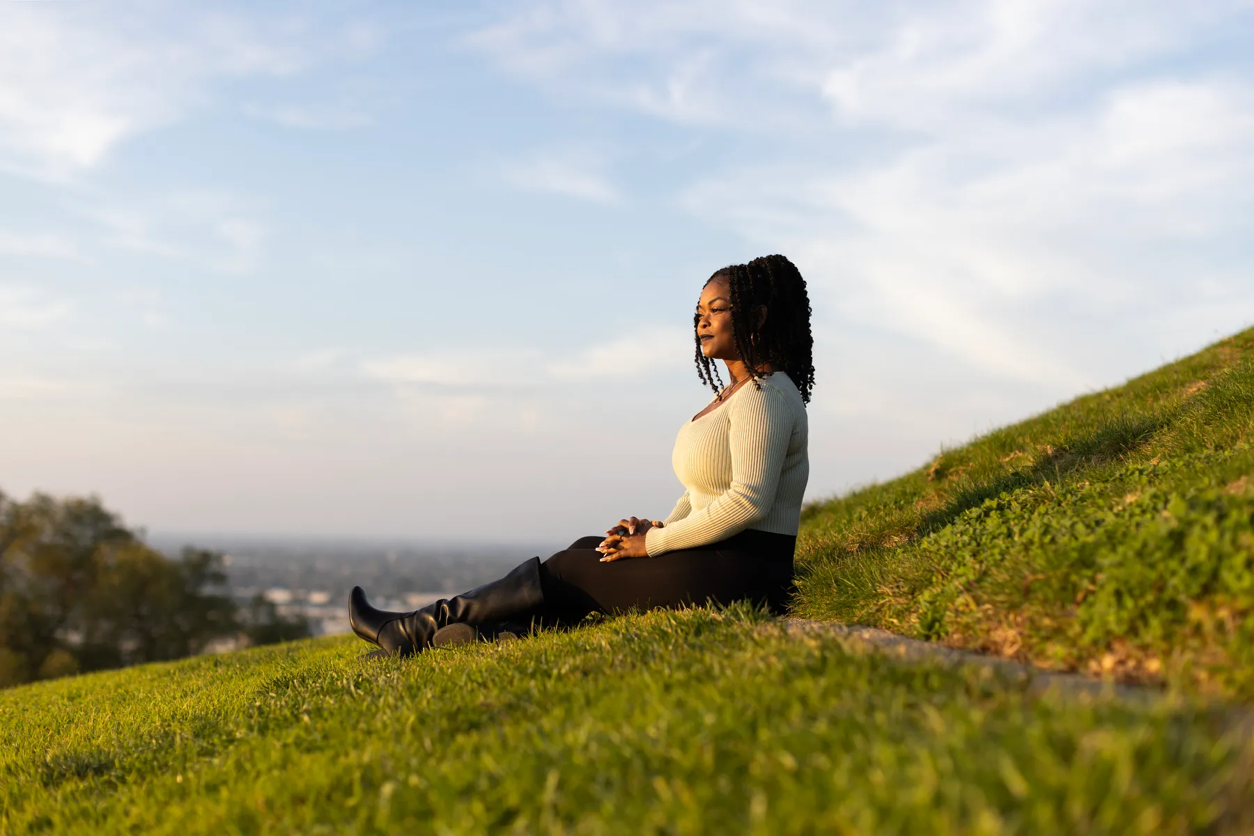 A woman sits on a grassy hill with the sky behind her.