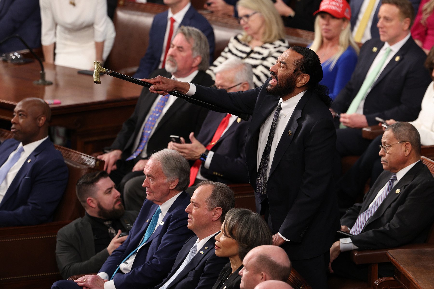 A man stands up in a room of seated politicians, pointing his cane at an unseen person while he shouts.