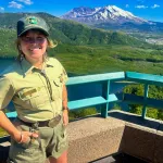 A woman in a park ranger uniform smiles at the camera in front of a lake and mountains.