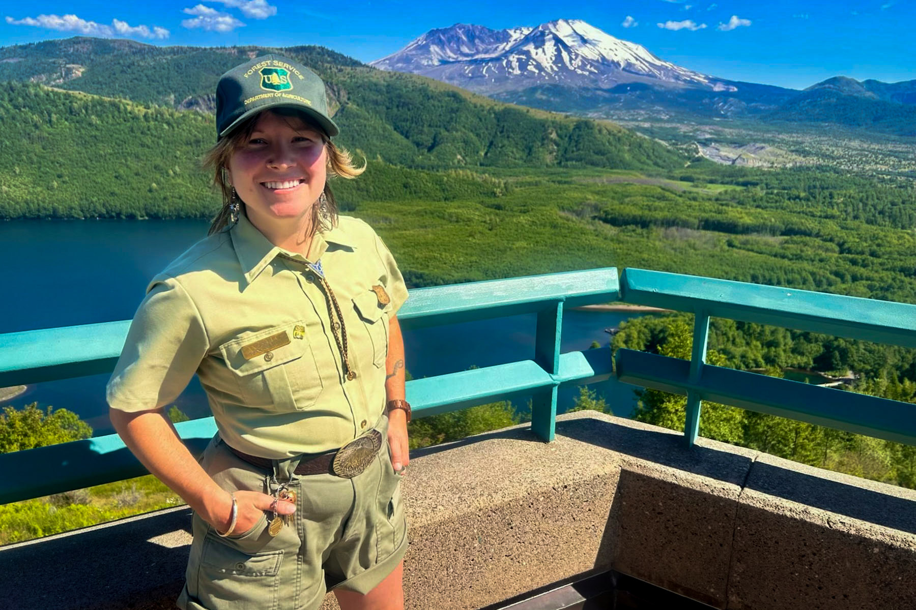 A woman in a park ranger uniform smiles at the camera in front of a lake and mountains.