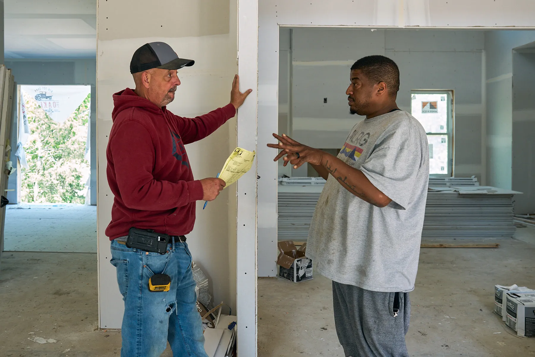Isa Conway talks with a man in a red hoodie and baseball cap inside an active construction site.