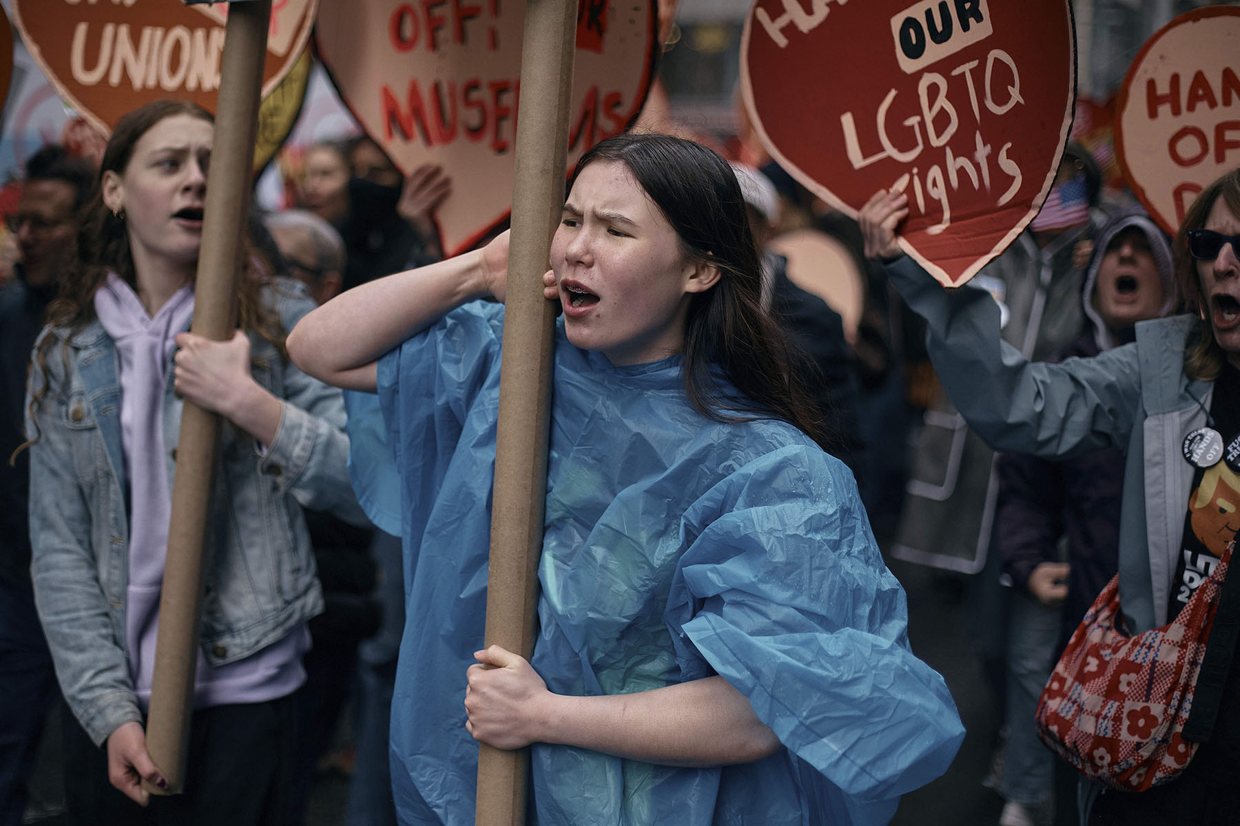 Uma jovem em um poncho de chuva azul grita durante um protesto na cidade de Nova York. Ela segura uma placa e é cercada por outros manifestantes que seguram sinais em forma de coração vermelho que leem slogans como 