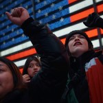 Protestors stand in front of a large digital American flag display in New York City.