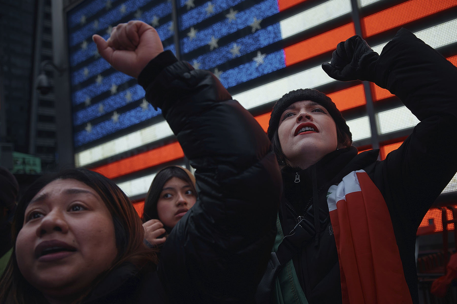 Protestors stand in front of a large digital American flag display in New York City.