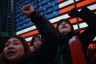 Protestors stand in front of a large digital American flag display in New York City.