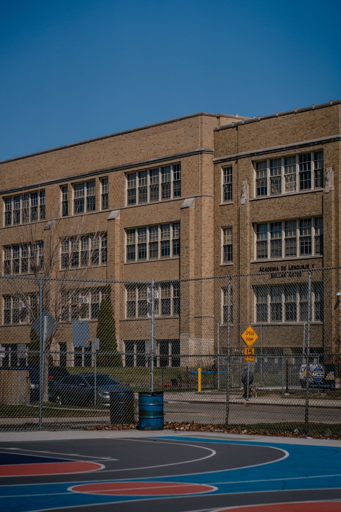 A tan brick school building behind a chain-link fence and a basketball court.