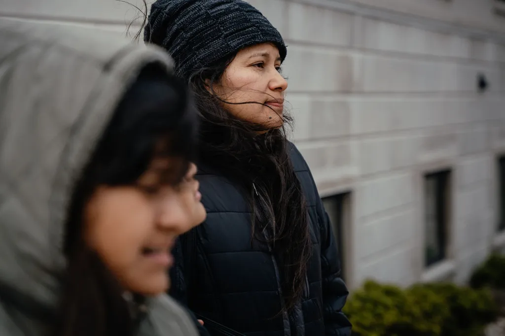 Yessenia Ruano is framed in focus while one daughter appears blurred in the foreground; both are wearing jackets and walking beside a government building.