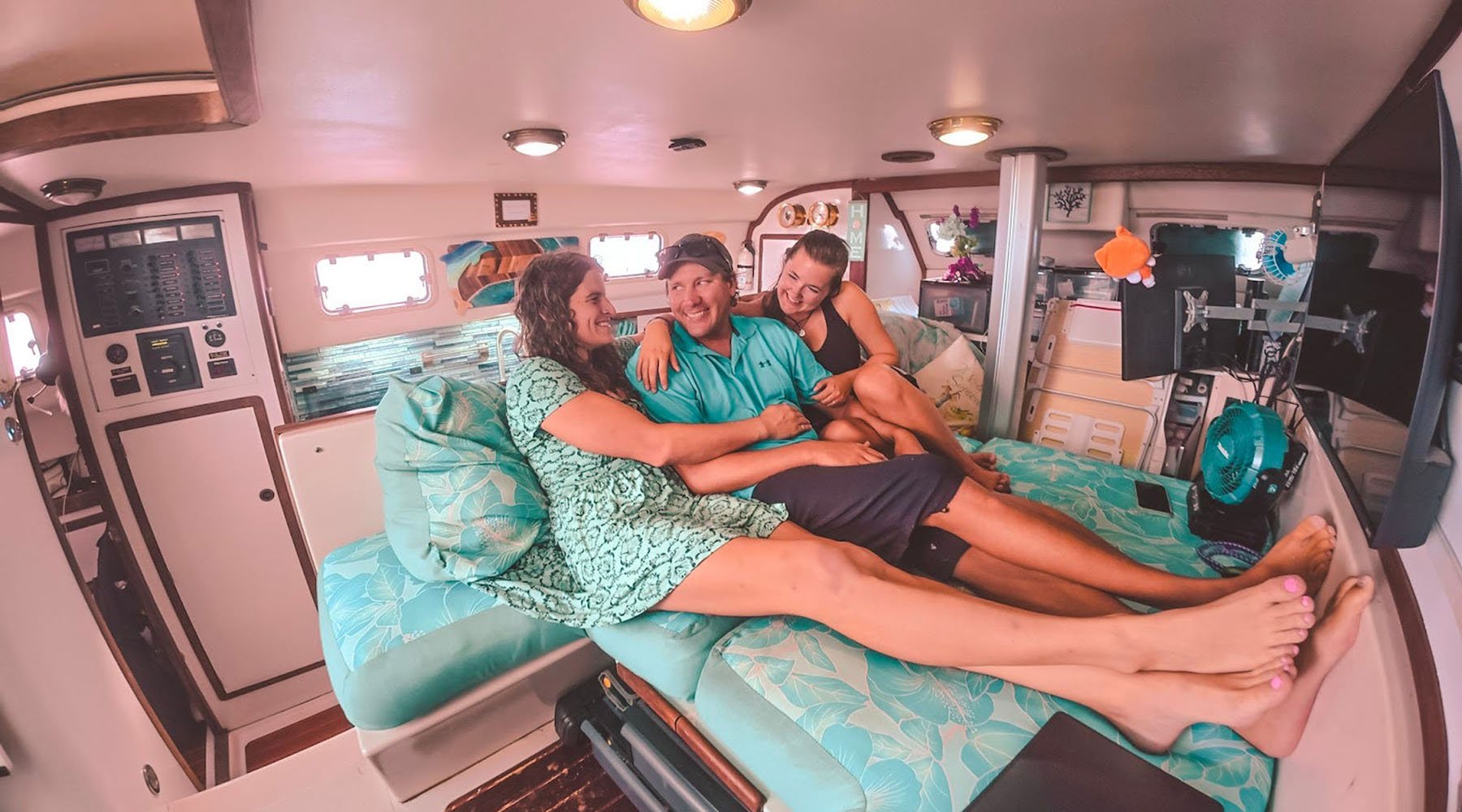 Three people lounge and laugh together on a teal-cushioned bed inside a boat cabin.
