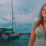 Kelsey Granger smiles while leaning against a mast with wind-blown hair. Behind her, a sailboat floats.