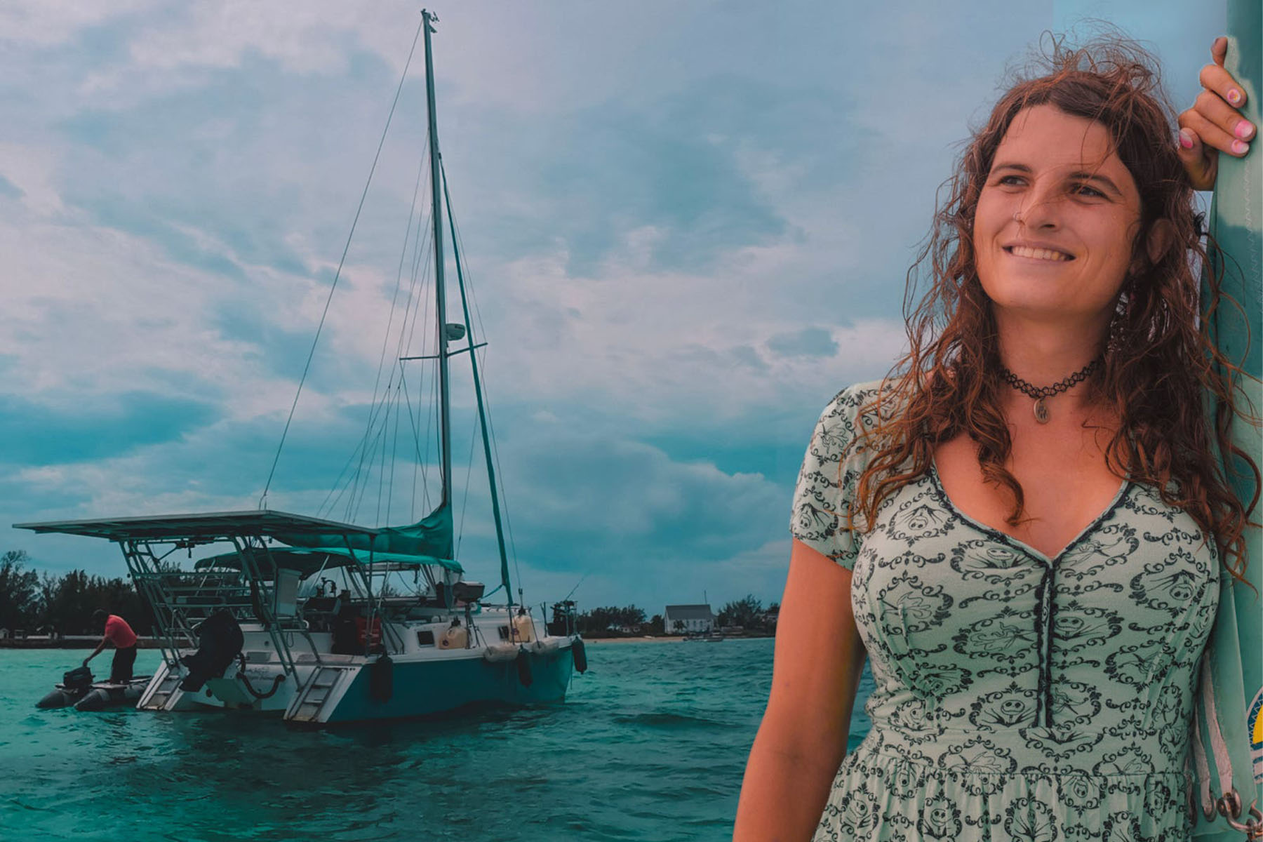 Kelsey Granger smiles while leaning against a mast with wind-blown hair. Behind her, a sailboat floats.