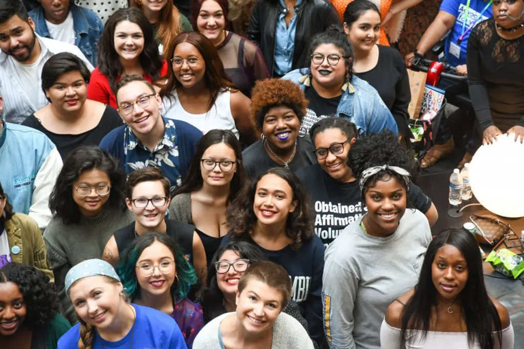 A large, diverse group of young people smiles up at the camera in a brightly lit indoor setting.