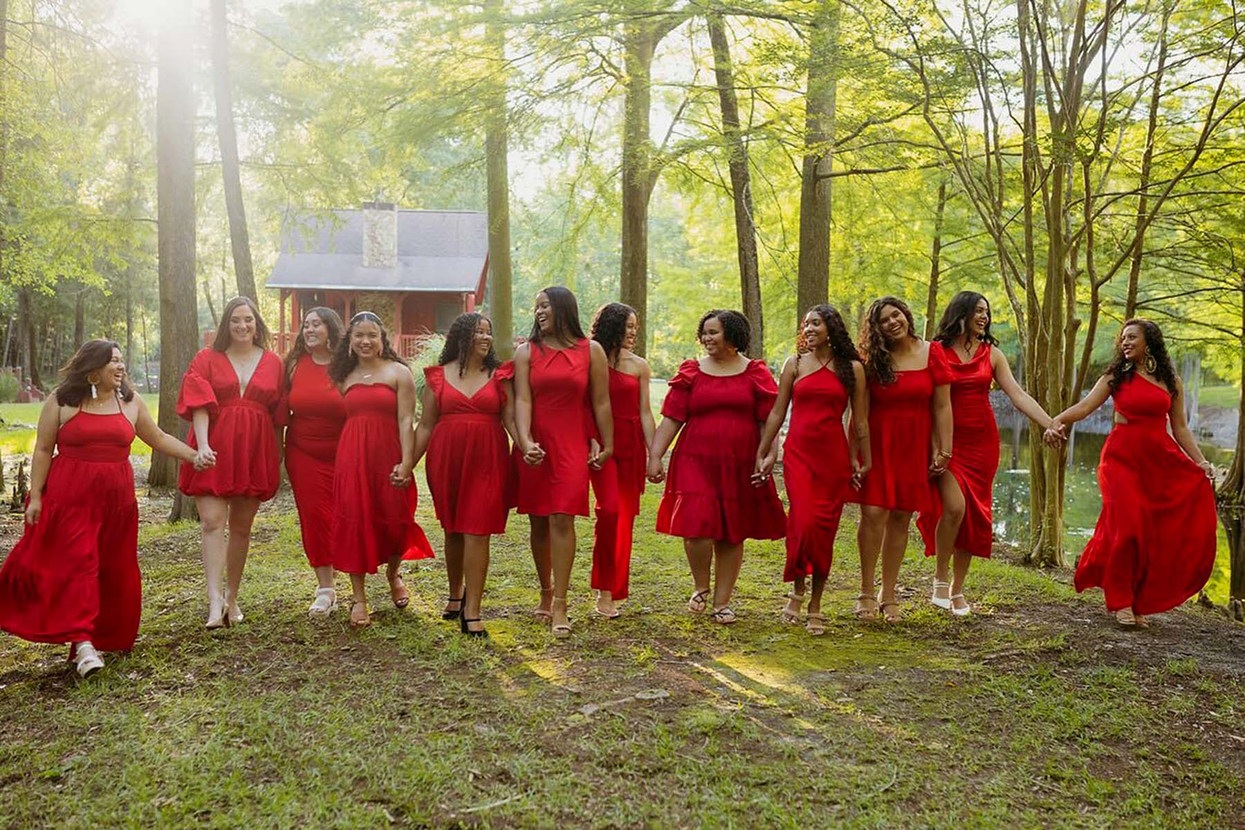 Twelve members of Alpha Pi Omega sorority, all wearing red dresses, walk hand in hand through a wooded area near a cabin.