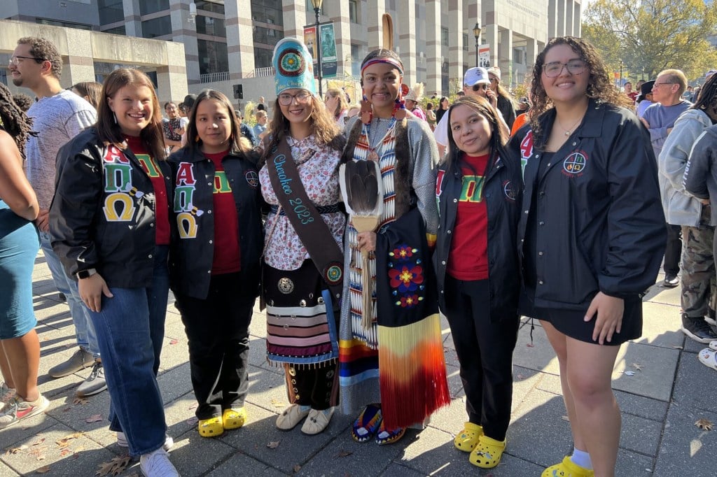 Six women from the Alpha Pi Omega sorority pose together at an outdoor gathering in Raleigh, North Carolina. Five of them wear matching black Greek letter jackets and red shirts; two women at the center wear traditional Indigenous regalia.
