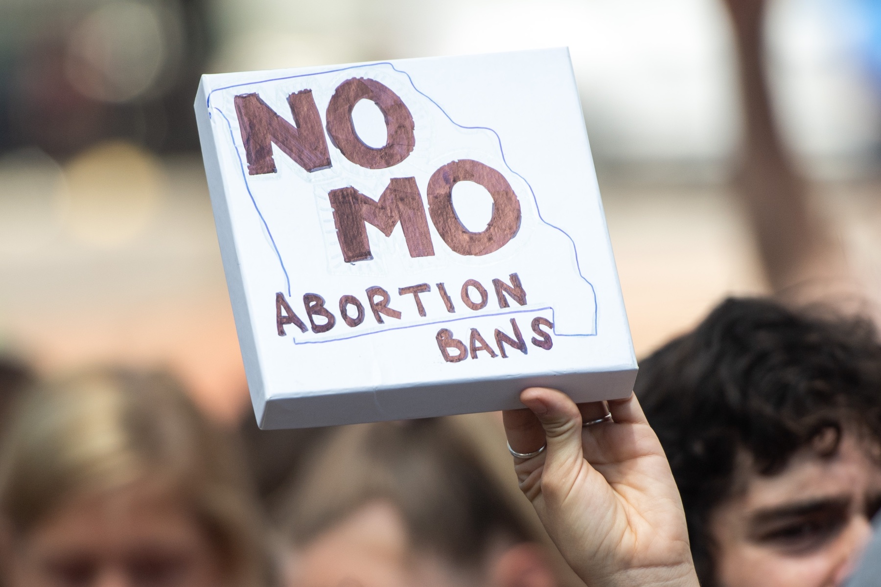 A hand holding a small white sign that reads "NO MO ABORTION BANS" with the state of Missouri drawn in outline.