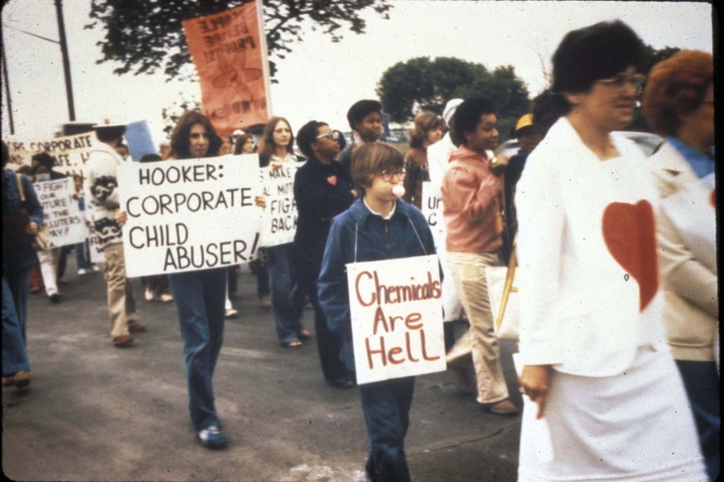 Residents of Love Canal march in protest, holding signs that read “Hooker: Corporate Child Abuser” and “Chemicals Are Hell.” The protest took place on Mother’s Day around 1980.